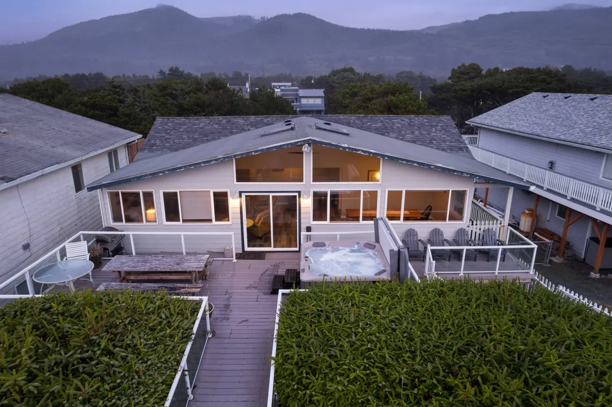 The image showcases a spacious oceanfront deck with a bubbling hot tub, ample seating, and a fire pit, all overlooking sweeping mountain and beach views. The serene patio invites guests to unwind, watch sunsets, or stargaze under the coastal sky, evoking a tranquil and luxurious retreat vibe.