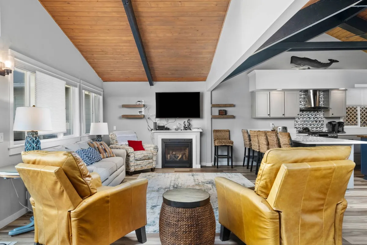 A welcoming great room with a central fireplace, a large TV, plush seating, and a kitchen area visible in the background. The room features expansive windows that let in natural light, giving glimpses of the ocean view and beach grasses outside. The open-plan layout connects the living, dining, and kitchen spaces seamlessly.