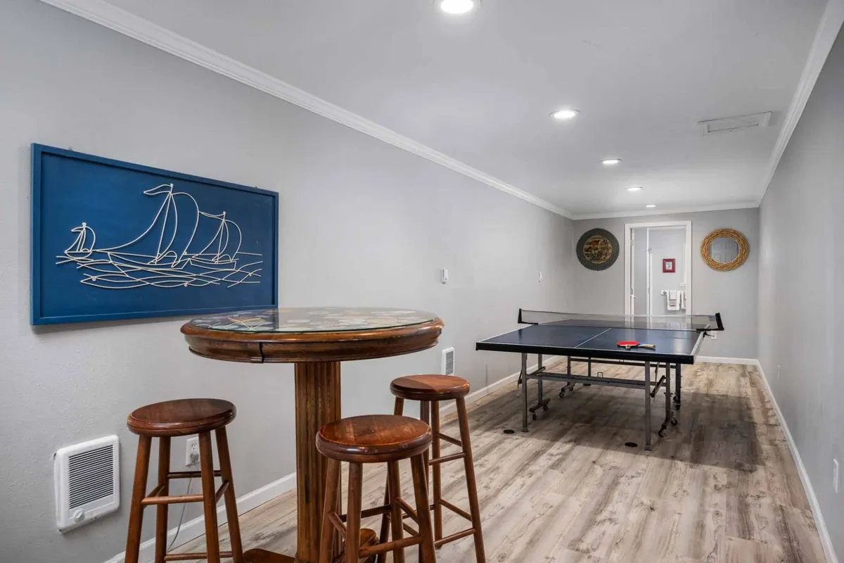A bright, engaging game room centered around a ping pong table, with bar stools for spectators or casual conversation, and cheerful sailboat wall art. The area invites lively competition and camaraderie.