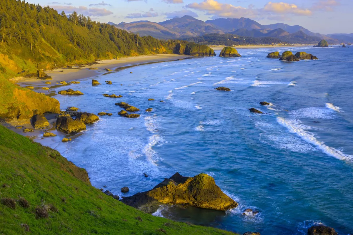 An expansive ocean vista featuring flowing waves, dramatic rock formations, and a backdrop of distant mountains. The scene is serene, highlighting the natural beauty and spaciousness offered by the location.