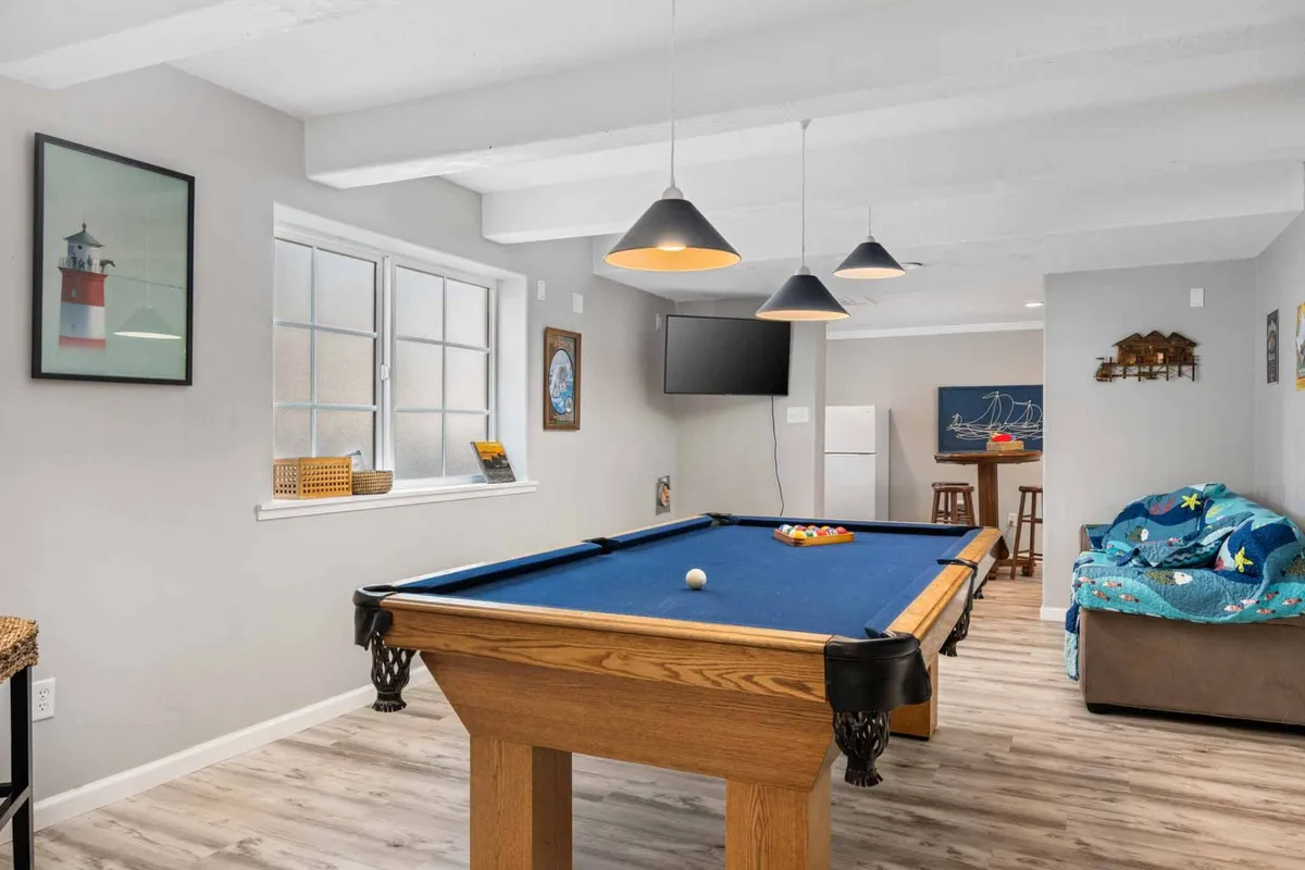 This image showcases a spacious, light-filled living area featuring a comfortable sofa positioned near floor-to-ceiling windows, a pool table for entertainment, modern hanging light fixtures, and elegant wall art. The arrangement subtly hints at coastal views just outside the glass, with an inviting space perfect for relaxation or socializing.