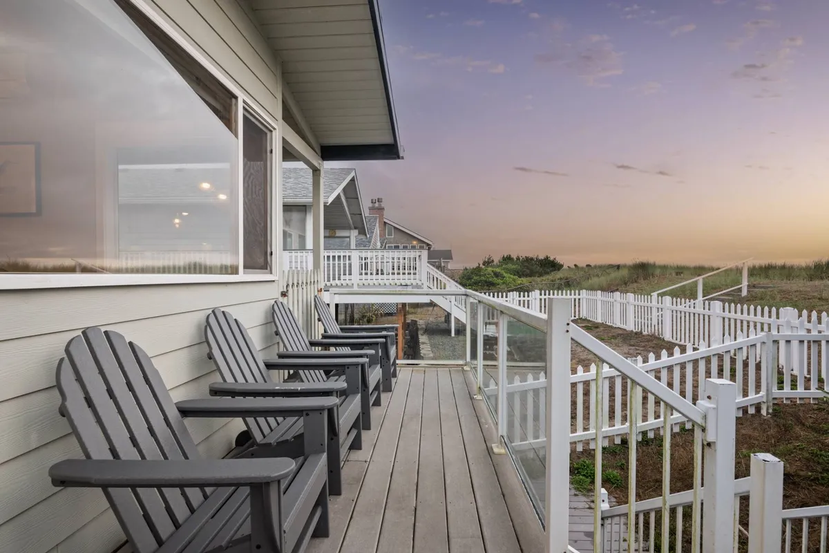 A tranquil exterior view of the home's wooden deck featuring adirondack chairs and a white picket fence, with the horizon glowing at sunset. The setup invites guests to gather, unwind, and soak in the ocean ambiance together.