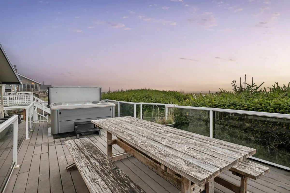 The image showcases the home's expansive deck featuring a bubbling hot tub, a wooden picnic table, lush greenery, and a sky painted by sunset. The setting is serene, ideal for unwinding after a day of exploration or hosting a family BBQ with ocean views.
