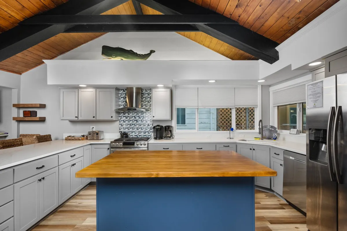 The image highlights a cheerful kitchen scene featuring a prominent island, blue cabinetry, exposed wooden beams, and a decorative backsplash. The layout encourages interaction and togetherness, with ample workspace for multiple cooks and a welcoming vibe for shared meals or cooking lessons.