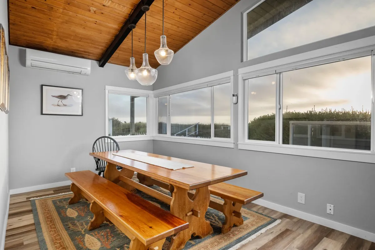 A striking dining area with a large wooden table and hanging light fixtures, positioned beside windows that frame ocean or beach views. The setting is warm and communal, perfect for shared meals and conversation with scenic backdrops.