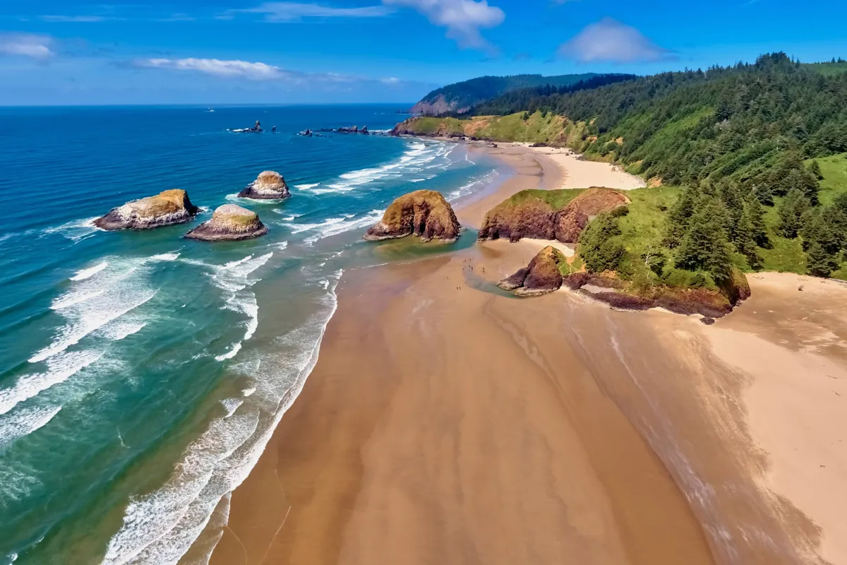 A breathtaking exterior view of rock formations, sandy beach, rolling waves, and green hills—highlighting the natural beauty of the nearby coastline. The scenery invites beachcombing, sunset picnics, and group exploration.